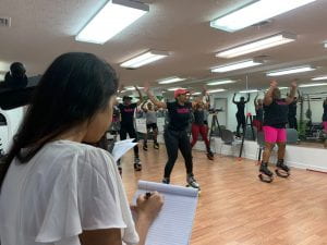 A group fitness class exercises using jump boots while an MBA student takes notes on the side.