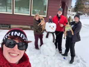 Chris and her family stand in front of a snowman with their two pug dogs..
