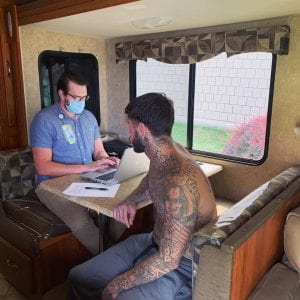 A provider wearing scrubs sits a a table inside a mobile clinic and registers a patient wearing no shirt with tattoos.