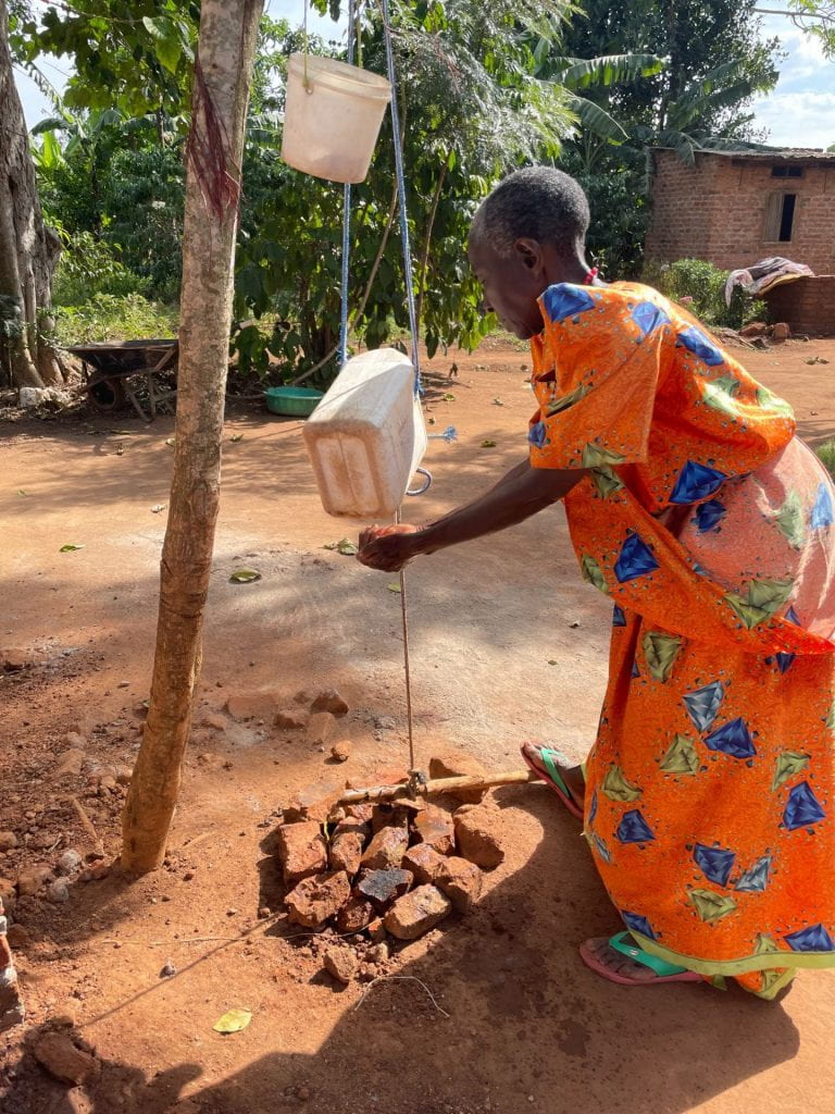 A woman washes her hands under a white plastic jug of water hanging from a tree.