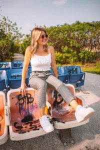 A woman models Everewear clothing sitting on graffitied seats: white tank top and grey pants.